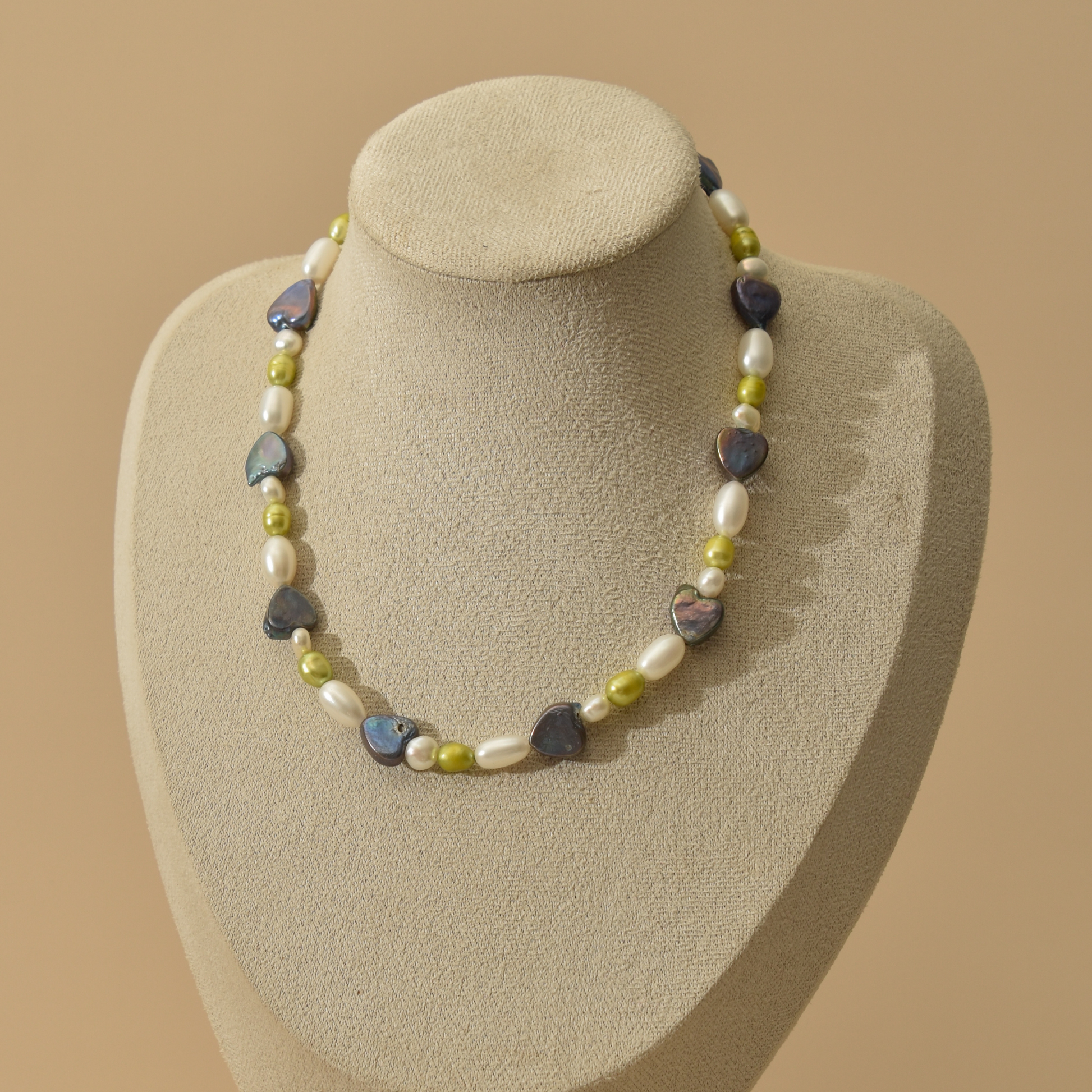A pearl necklace displayed on a beige jewelry bust against a warm beige background. The necklace features irregular dark blue pearls, white baroque pearls, and small green pearls.