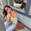 A woman wearing a grey and yellow striped off-the-shoulder top and light blue jeans sits on a sidewalk next to a building, with a brown leather handbag resting beside her.