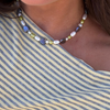 A close-up of a person wearing a necklace with white pearls, blue heart pearls, and olive-green pearls, paired with a blue and cream striped top.