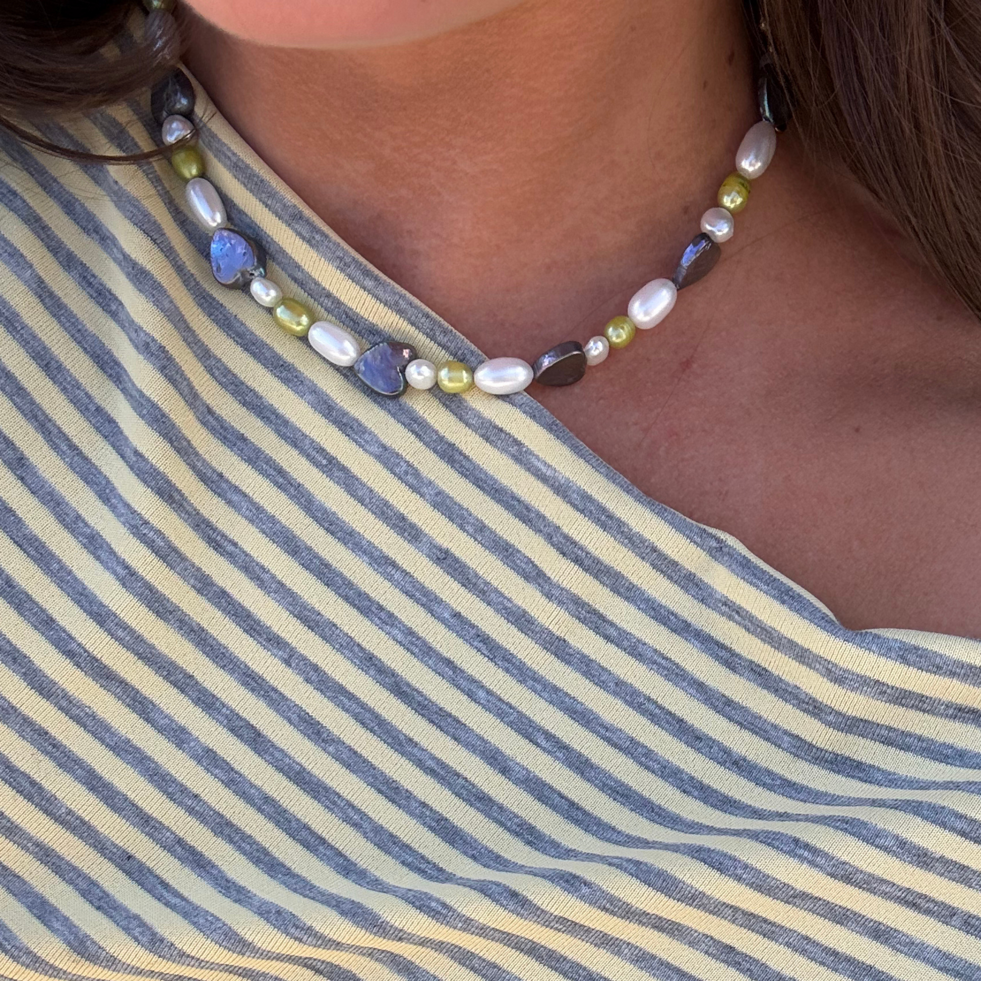 A close-up of a person wearing a necklace with white pearls, blue heart pearls, and olive-green pearls, paired with a blue and cream striped top.