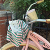 A pink vintage-style bicycle with a green and pink striped fabric bag hanging from the front, alongside a white tote bag and a pearl necklace, set against a background of green foliage and a brick wall.