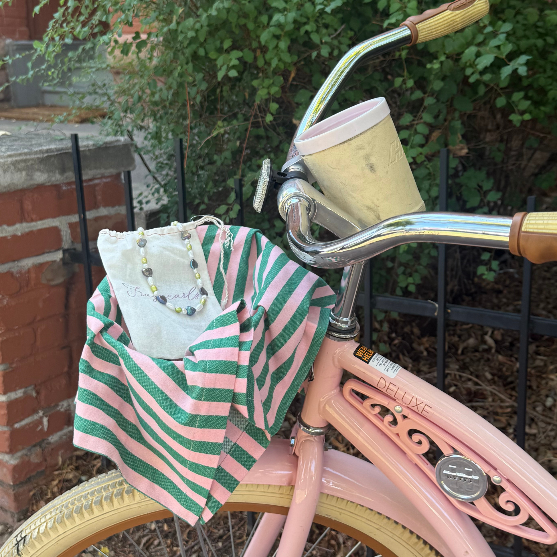 A pink vintage-style bicycle with a green and pink striped fabric bag hanging from the front, alongside a white tote bag and a pearl necklace, set against a background of green foliage and a brick wall.