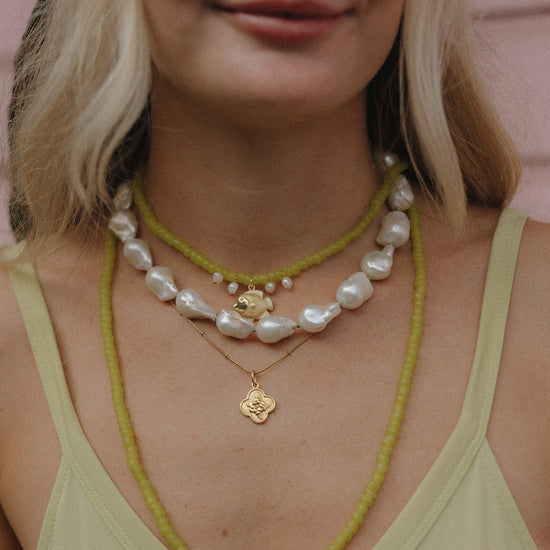 A woman wears layered necklaces: green beads, large irregular pearls, and gold pendants on a light green top. The style is casual and elegant.