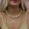 A woman wears layered necklaces: green beads, large irregular pearls, and gold pendants on a light green top. The style is casual and elegant.