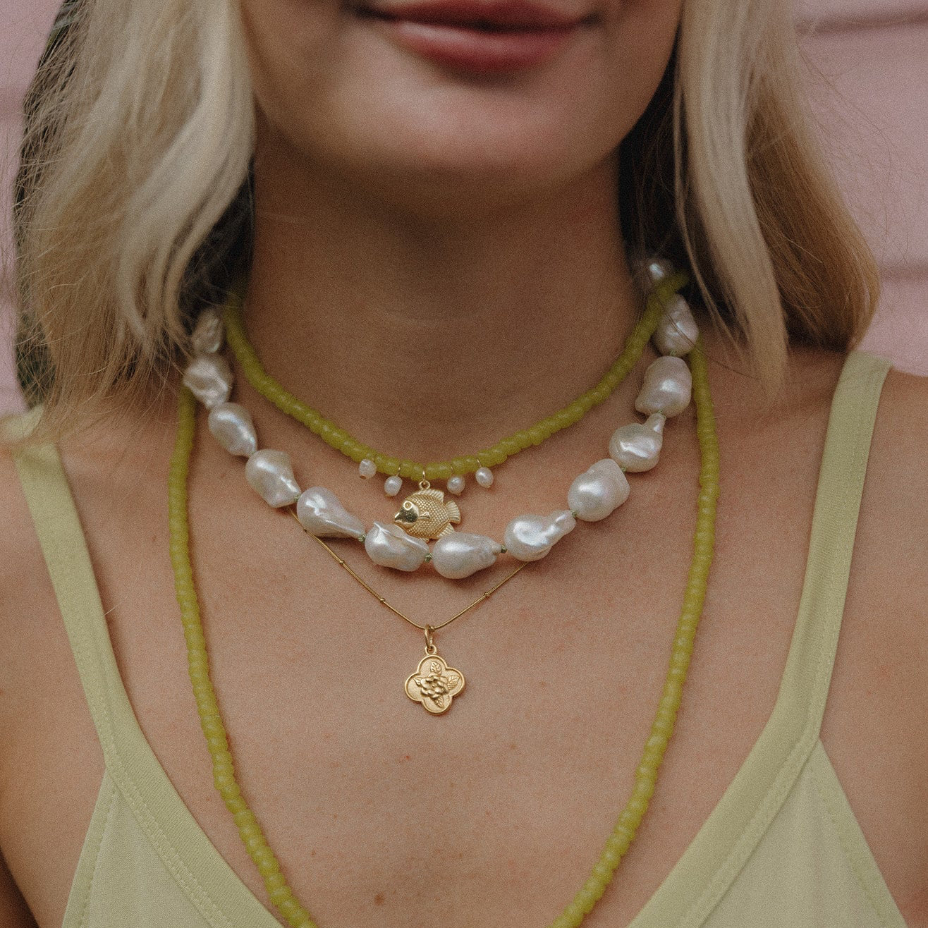 A woman wears layered necklaces: green beads, large irregular pearls, and gold pendants on a light green top. The style is casual and elegant.