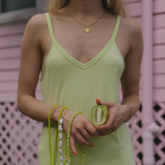 A person in a light green dress holds a sliced kiwi in one hand, with the other hand adorned with lime green beads, pearls, and gold bracelets.