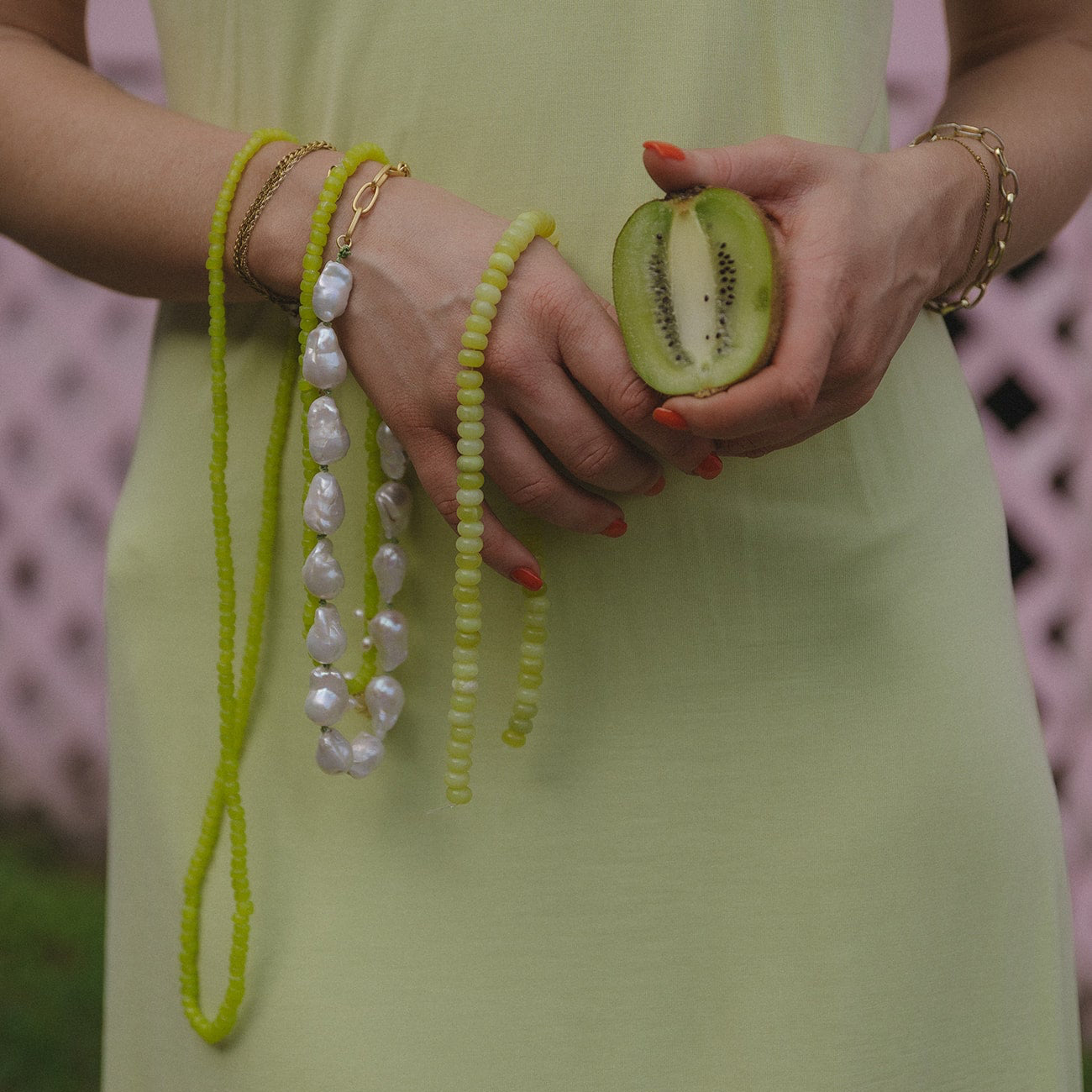 A person in a light green dress holds a sliced kiwi in one hand, with the other hand adorned with lime green beads, pearls, and gold bracelets.