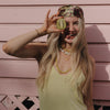 A woman playfully holds a kiwi slice over one eye, sticking out her tongue. She wears a patterned headscarf, necklaces, and a light tank top. The background is pink siding, conveying a fun and carefree mood.