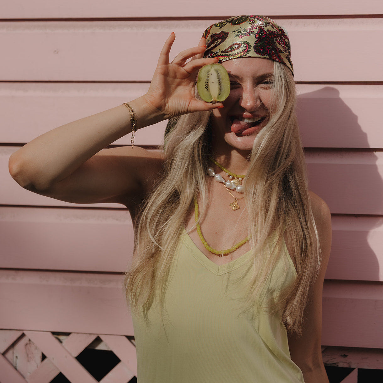 A woman playfully holds a kiwi slice over one eye, sticking out her tongue. She wears a patterned headscarf, necklaces, and a light tank top. The background is pink siding, conveying a fun and carefree mood.