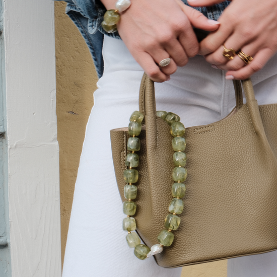 Close-up of a person holding a beige textured handbag with a green beaded strap. They wear white pants, a denim jacket, rings, and a bracelet. The mood is casual and stylish.