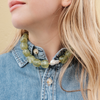 A woman with long blonde hair wears a denim shirt and a bold green stone necklace with pearls. Her expression is calm, set against a beige background.