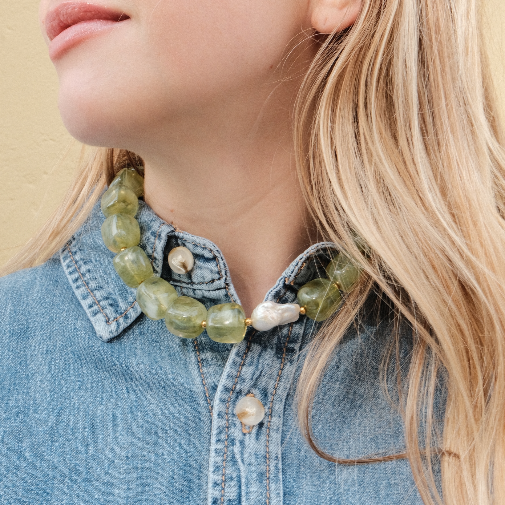 A woman with long blonde hair wears a denim shirt and a bold green stone necklace with pearls. Her expression is calm, set against a beige background.