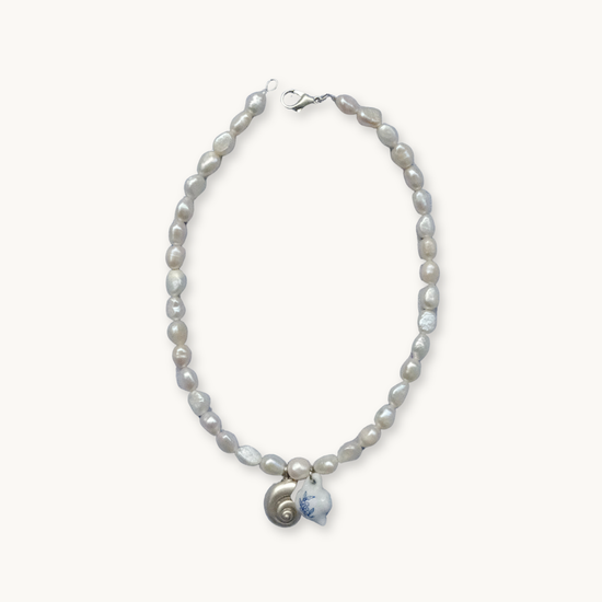A necklace with irregular pearls features a metal clasp. The centerpiece includes a small spiral shell and a white pendant with blue detailing, exuding an elegant, coastal vibe.