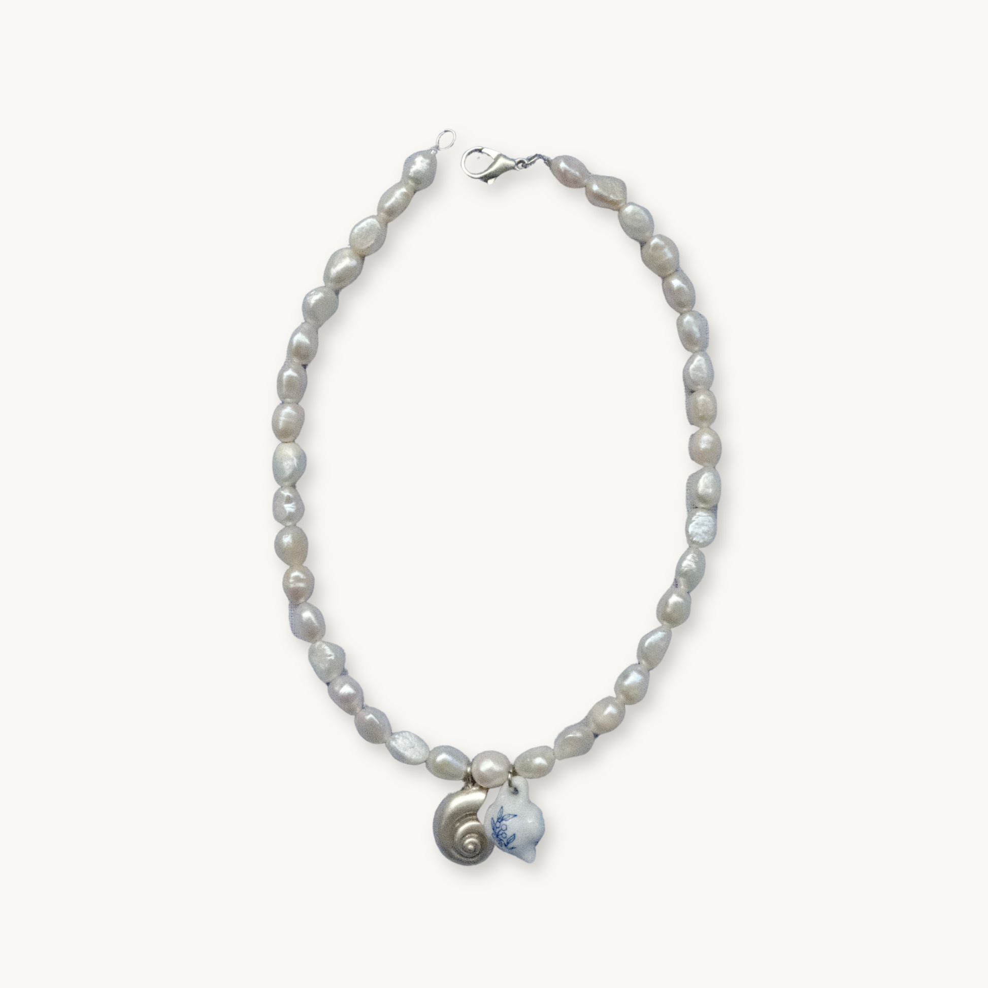 A necklace with irregular pearls features a metal clasp. The centerpiece includes a small spiral shell and a white pendant with blue detailing, exuding an elegant, coastal vibe.