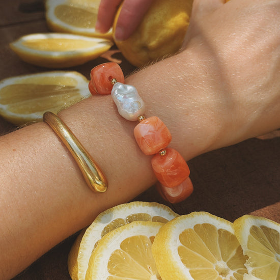 A wrist adorned with vibrant orange and pearl beads, alongside a sleek gold bracelet. Sliced lemons surround the scene, creating a fresh, summery vibe.