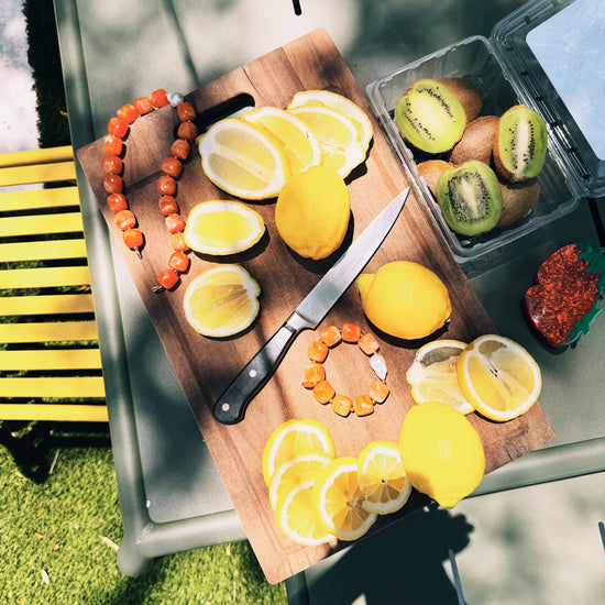 Slices of bright yellow lemons and whole lemons are arranged on a wooden cutting board with a knife. A bracelet made of orange beads lies alongside. Nearby, whole and halved kiwis in a plastic container add a touch of green, all bathed in warm sunlight, creating a fresh, vibrant atmosphere.