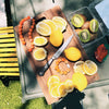 Slices of bright yellow lemons and whole lemons are arranged on a wooden cutting board with a knife. A bracelet made of orange beads lies alongside. Nearby, whole and halved kiwis in a plastic container add a touch of green, all bathed in warm sunlight, creating a fresh, vibrant atmosphere.