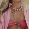 A person in a pink outfit wears layered necklaces with pearls and beads, featuring gold and green pendants. The tone is relaxed and summery.
