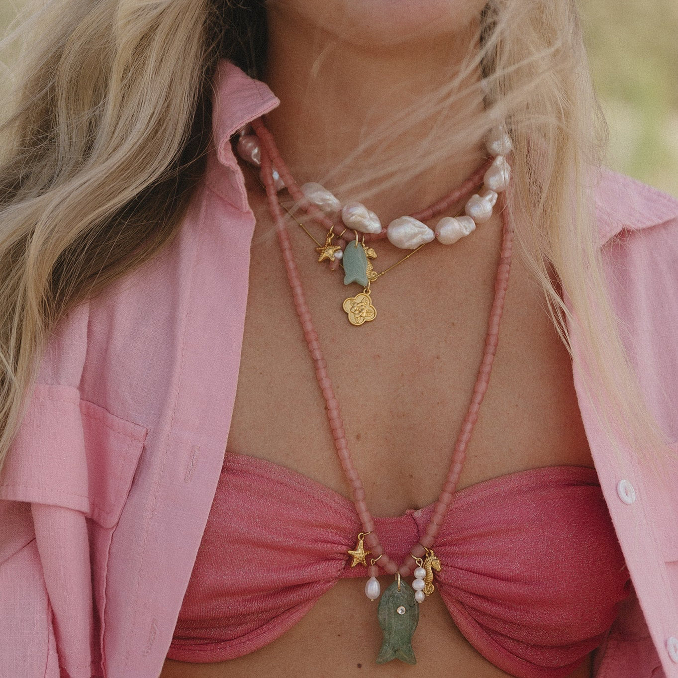 A person in a pink outfit wears layered necklaces with pearls and beads, featuring gold and green pendants. The tone is relaxed and summery.