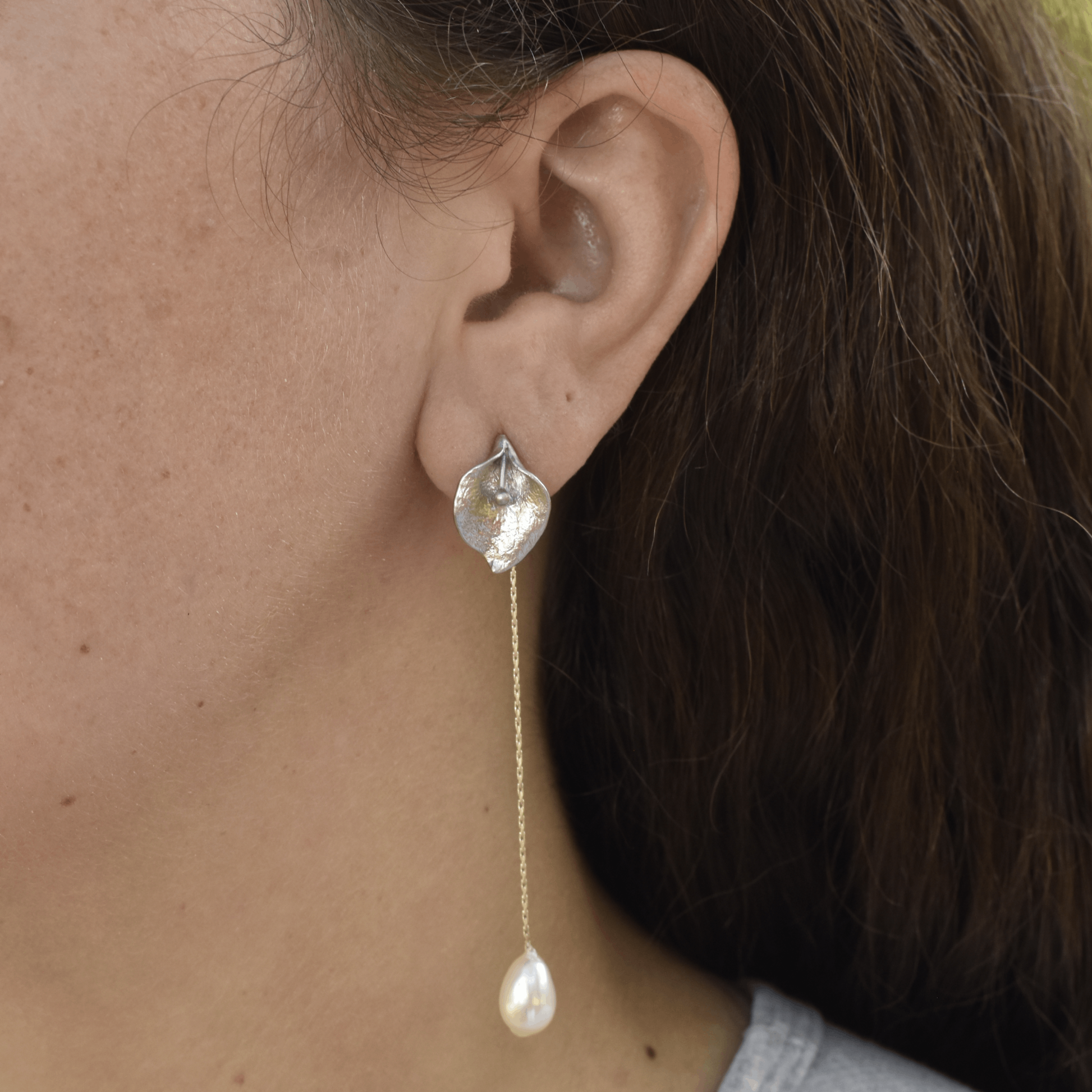A close-up of a person's ear wearing an elegant earring featuring a silver petal design with a long gold chain and a teardrop pearl at the end.