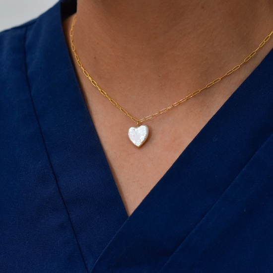 A gold chain necklace with a heart-shaped pearl pendant is worn around a person's neck, set against a blue collared shirt background.