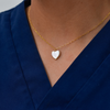 A gold chain necklace with a heart-shaped pearl pendant is worn around a person's neck, set against a blue collared shirt background.