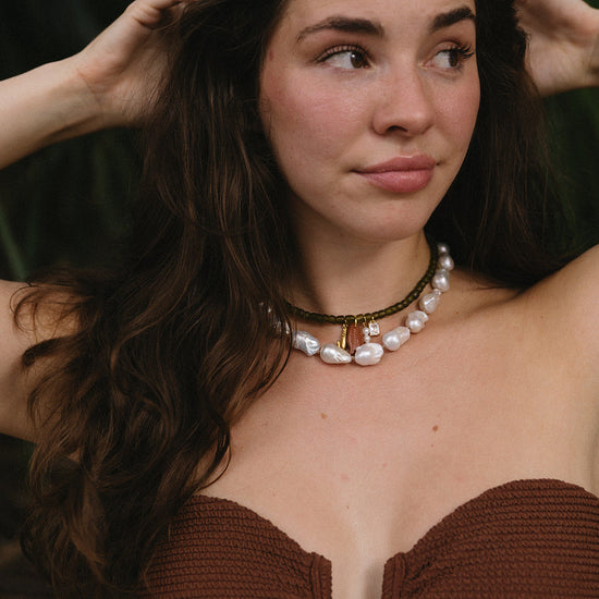 Woman with long brown hair wearing a strapless textured top and layered necklaces with pearls and charms. She gazes thoughtfully to the side.