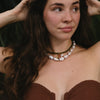 Woman with long brown hair wearing a strapless textured top and layered necklaces with pearls and charms. She gazes thoughtfully to the side.