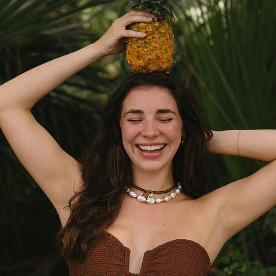 A woman with long dark hair smiles with pearl necklaces while holding a pineapple above her head. She's in a tropical setting with lush green palm leaves.