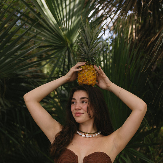 A woman with long dark hair and pearl necklace smiles while holding a pineapple above her head. She's in a tropical setting with lush green palm leaves.