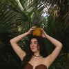 A woman with long dark hair and pearl necklace smiles while holding a pineapple above her head. She's in a tropical setting with lush green palm leaves.