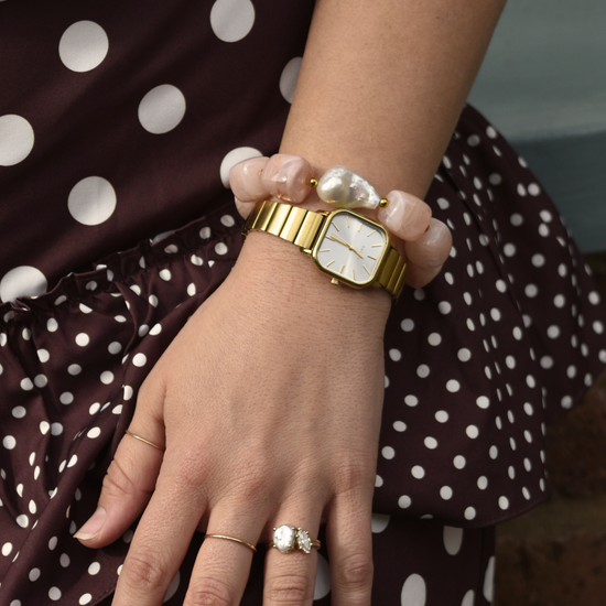 Close-up of a wrist wearing a gold watch with a white square face and a chunky pink beaded bracelet. Background shows a maroon polka dot fabric.