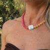 A woman wears a colorful necklace with pink beads on one side, yellow on the other, and a central white, iridescent pearl. The setting is bright and natural.
