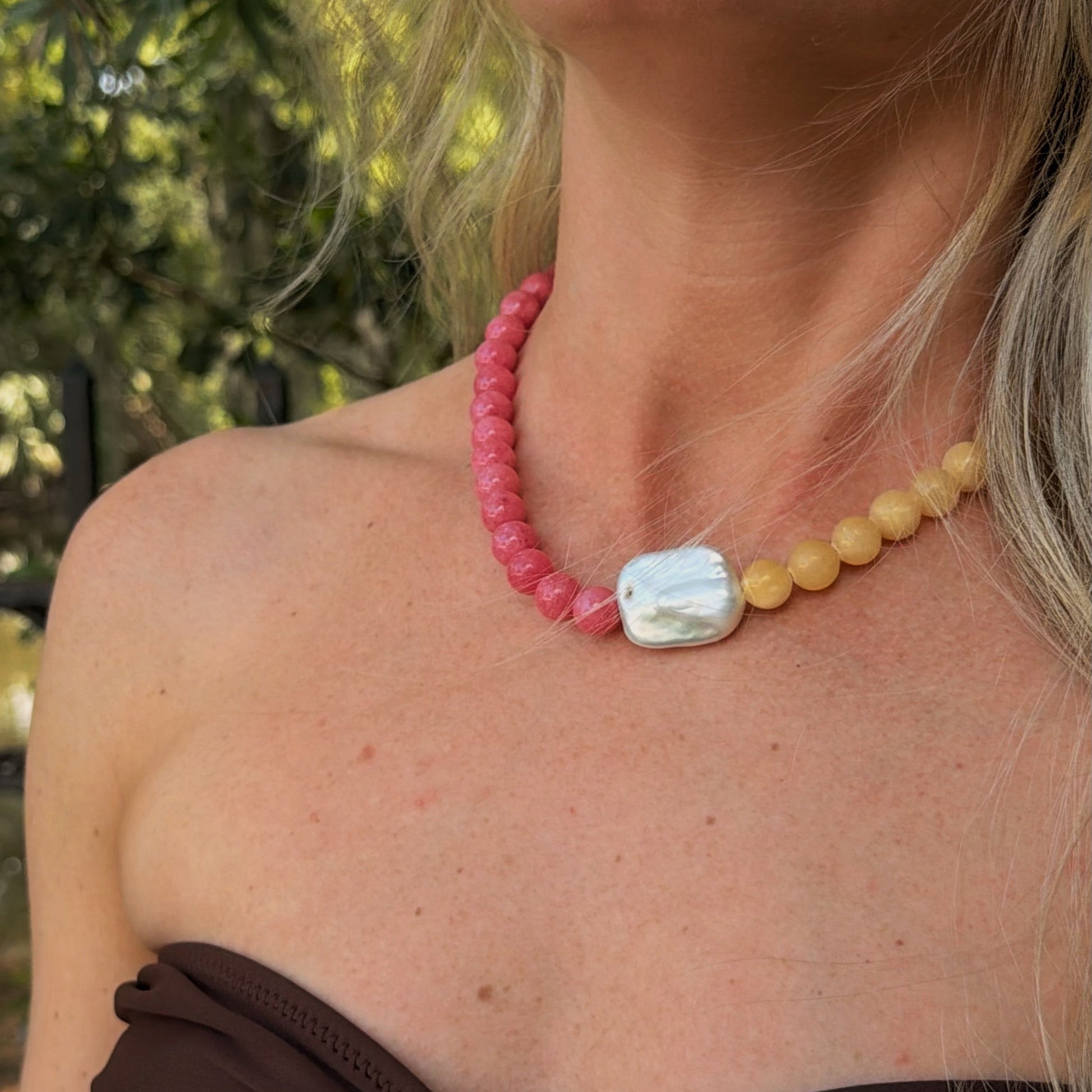 A woman wears a colorful necklace with pink beads on one side, yellow on the other, and a central white, iridescent pearl. The setting is bright and natural.