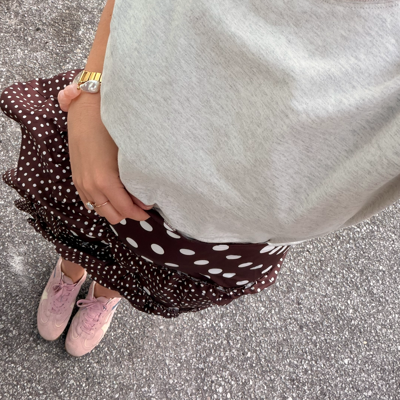 A person is standing on a pavement wearing a gray top, a brown polka-dot skirt, a pink stone and pearl bracelet, and pink sneakers. Their hand rests on their hip.