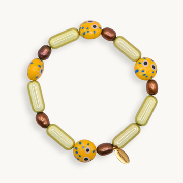 A handmade beaded bracelet with olive-green capsule beads, yellow-orange round beads with blue and black speckles, and metallic bronze oval beads, displayed on a light cream background.