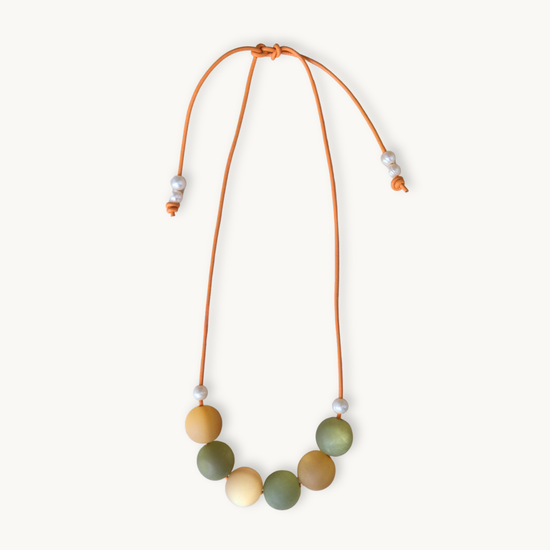 A beaded necklace with a flexible orange cord, featuring seven matte-colored round beads in shades of olive green, mustard yellow, and cream, accented with small white spacer beads, shown on a light cream background.