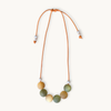 A beaded necklace with a flexible orange cord, featuring seven matte-colored round beads in shades of olive green, mustard yellow, and cream, accented with small white spacer beads, shown on a light cream background.