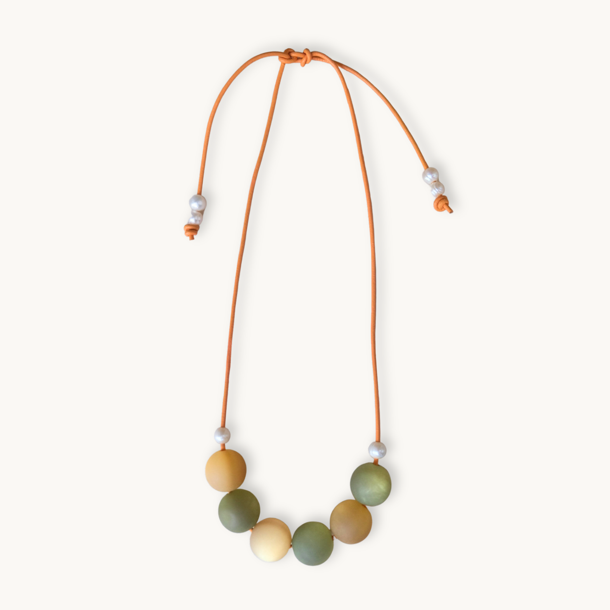 A beaded necklace with a flexible orange cord, featuring seven matte-colored round beads in shades of olive green, mustard yellow, and cream, accented with small white spacer beads, shown on a light cream background.