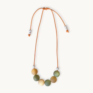 A beaded necklace with a flexible orange cord, featuring seven matte-colored round beads in shades of olive green, mustard yellow, and cream, accented with small white spacer beads, shown on a light cream background.
