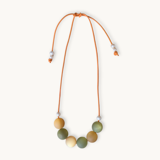 A beaded necklace with a flexible orange cord, featuring seven matte-colored round beads in shades of olive green, mustard yellow, and cream, accented with small white spacer beads, shown on a light cream background.
