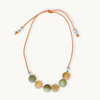 A beaded cord necklace featuring seven round, matte-colored beads in shades of sage green, peach, and light orange, suspended on an adjustable orange cord with small white spacer beads.
