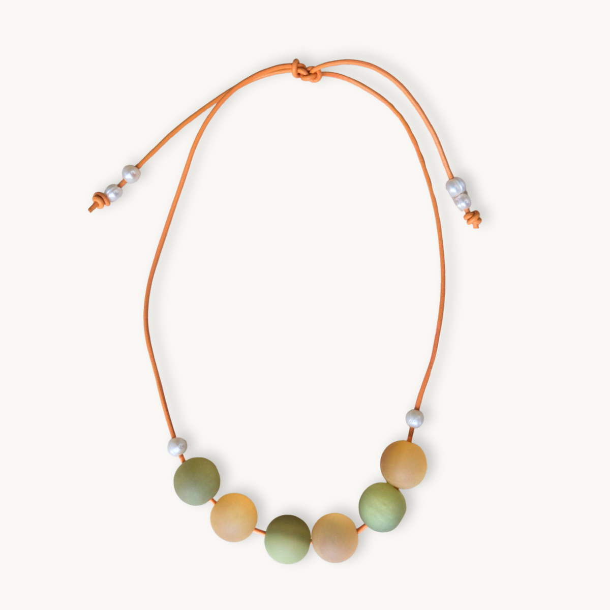 A beaded cord necklace featuring seven round, matte-colored beads in shades of sage green, peach, and light orange, suspended on an adjustable orange cord with small white spacer beads.