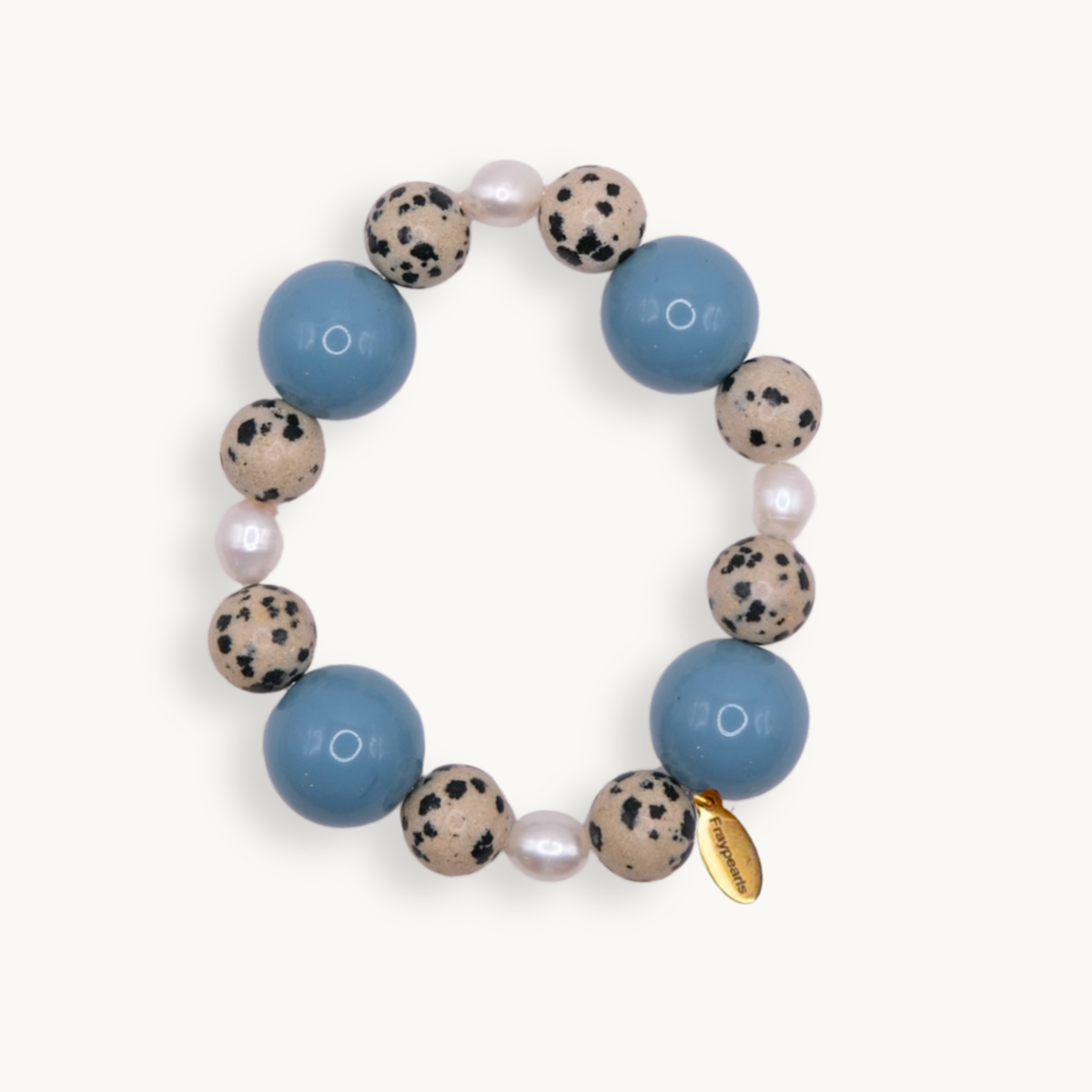 A beaded stretch bracelet featuring large light blue beads, speckled beige and black stone beads, small white pearls, and a small gold tag, shown on a light cream background.