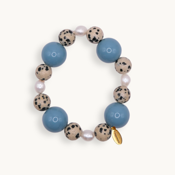 A beaded stretch bracelet featuring large light blue beads, speckled beige and black stone beads, small white pearls, and a small gold tag, shown on a light cream background.
