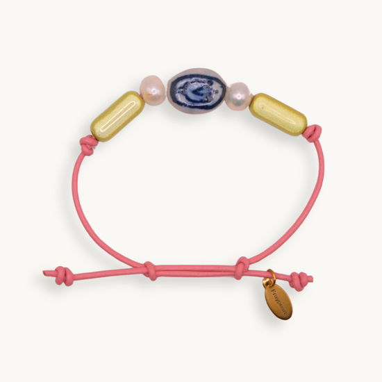 A pink cord bracelet with a sliding knot closure, decorated with a blue and white agate bead, two pink freshwater pearls, and two gold capsule beads. A small gold tag attached to the cord.