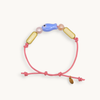 A pink cord bracelet featuring a small blue fish bead, two gold capsule beads, and two pink pearl-like beads, displayed on a light cream background.