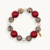 A beaded stretch bracelet featuring alternating red round beads, speckled beige and black stone beads, small white pearl-like beads, and a small gold tag, shown on a light cream background.