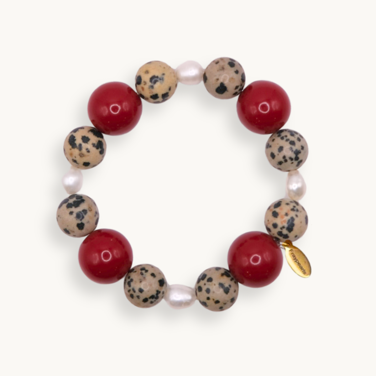 A beaded stretch bracelet featuring alternating red round beads, speckled beige and black stone beads, small white pearl-like beads, and a small gold tag, shown on a light cream background.