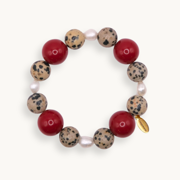 A beaded stretch bracelet featuring alternating red round beads, speckled beige and black stone beads, small white pearl-like beads, and a small gold tag, shown on a light cream background.